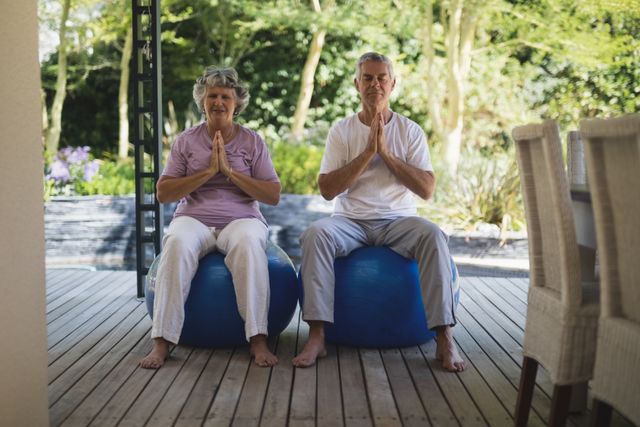 Senior couple practicing meditation on exercise balls at porch. Ideal for promoting healthy lifestyle, mindfulness, and wellness among elderly. Suitable for use in health and fitness blogs, retirement community advertisements, and wellness programs.