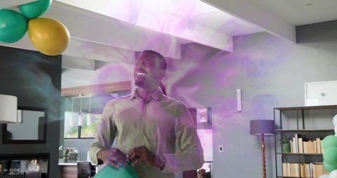 Smiling man holding green balloon celebrating in modern living room with purple haze