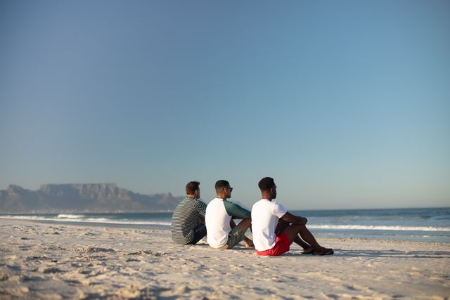 Three male friends are sitting on the sandy beach, enjoying a tranquil moment while looking at the ocean. The clear sky and distant mountains add to the scenic beauty. Ideal for use in travel promotions, friendship themes, leisure activities, and summer vacation advertisements.