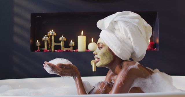 African american woman with towel and mask taking bath and blowing foam in bathroom. health and beauty concept.