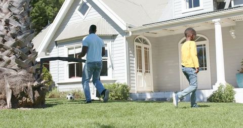 African american father and son carrying table across front lawn by porch with soccer ball