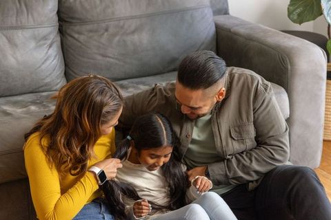 Multicultural family sitting and bonding while mother braiding daughter's hair by sofa