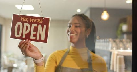 Smiling barista holding 'yes, we are open' sign welcoming customers to cozy coffee shop