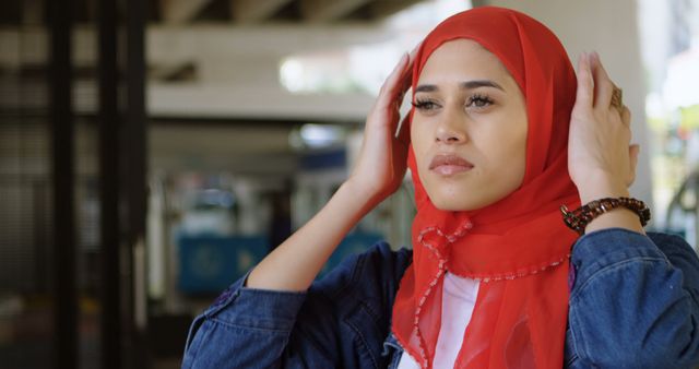 Young Muslim woman in traditional red hijab adjusting it with a serious expression while standing outdoors. The urban setting showcases modern life and cultural diversity. Useful for promoting inclusivity, cultural awareness, and discussions about traditional and contemporary lifestyles.