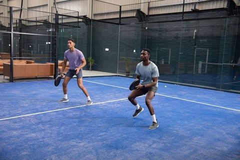 Two male athletes training doubles on blue indoor padel court with glass walls