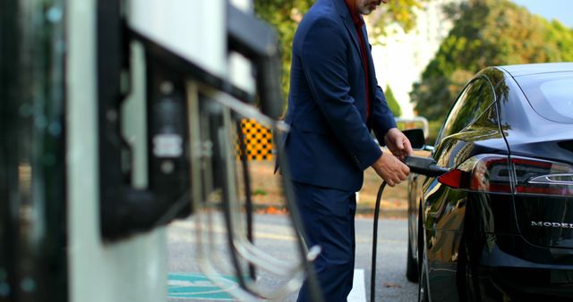 Businessman dressed in a blue suit plugs charger into electric car at outdoor charging station. Ideal for use in editorials about sustainable transportation, renewable energy solutions, and modern business practices. Can be utilized in green technology ads, corporate brochures, or articles focused on ecological advancements in mobility.