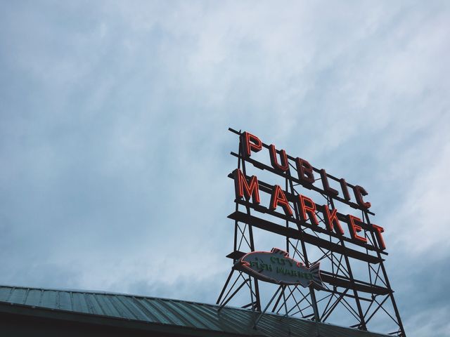 Capturing the iconic Public Market sign with an overcast sky above. The sign, featuring neon lights, stands out against the backdrop of cloud-filled skies, signifying a well-known urban landmark. Perfect for use in travel blogs, marketing materials for Seattle tourism, or historical archives highlighting prominent city landmarks.
