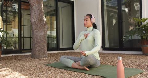 Meditating Asian woman practicing mindful breathing on green yoga mat outdoors with headphones
