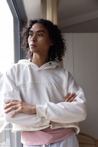 Non-binary standing by window looking out in white hoodie, contemplative home portrait