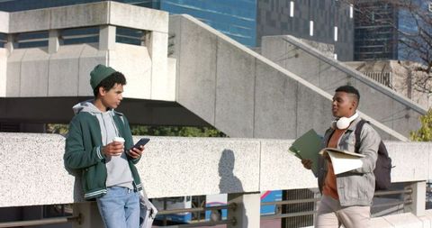 Urban college students chatting on brutalist campus walkway holding coffee, phone, folders