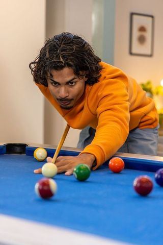 Young asian man lining up precise shot on blue-felt pool table in cozy living room
