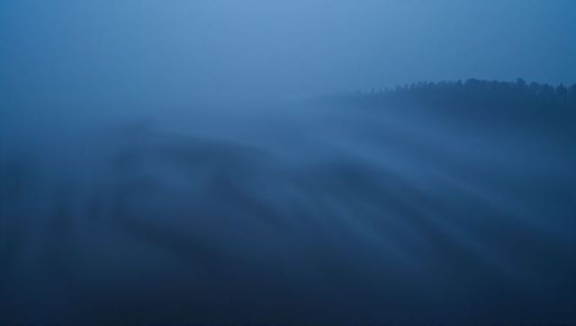 Rolling blue mist filling valley at pre-dawn, distant tree ridge silhouette, moody landscape
