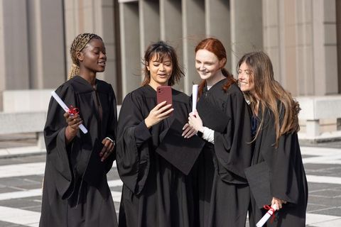 Diverse female graduates posing for selfie in black gowns on university plaza celebration