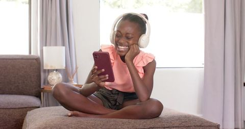 Joyful african teen listening to music on wireless headphones holding smartphone at home