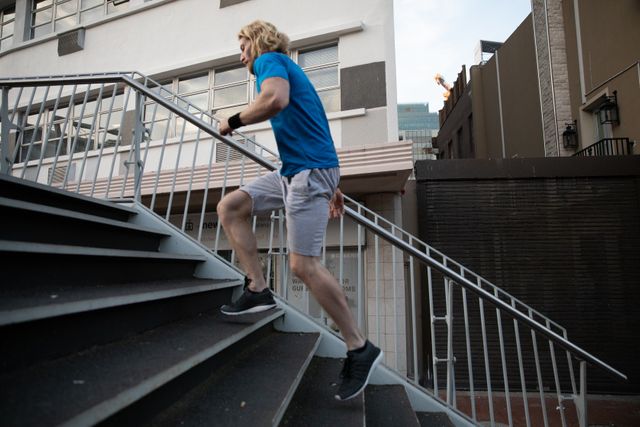Fit man with long blonde hair running upstairs in an urban environment. Ideal for promoting fitness, healthy lifestyle, outdoor workouts, and athletic apparel. Suitable for use in advertisements, fitness blogs, and health-related content.