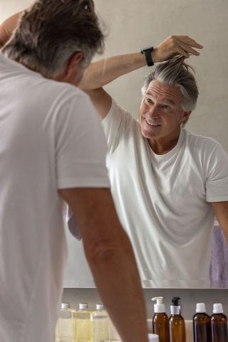 Senior man grooming at vanity mirror styling gray hair wearing smartwatch and smiling