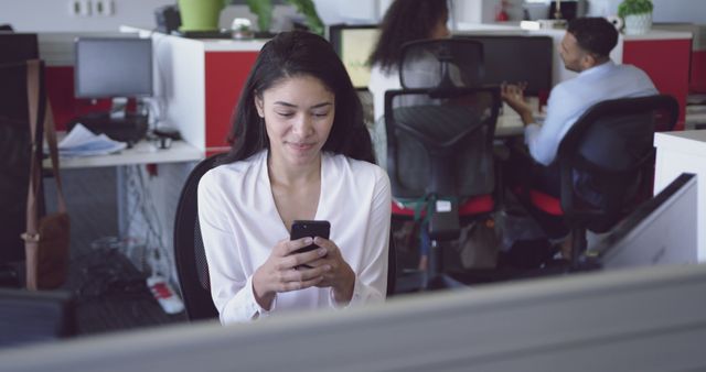 Biracial woman checks phone, peers work in background. She has long dark hair, wearing white blouse, in office