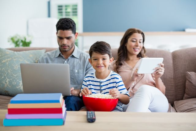 This image depicts a modern family in a relaxed home setting, with a father using a laptop, a mother using a tablet, and their young son eating popcorn while sitting between them. Useful for articles or ads related to family bonding, digital technology, work from home, parental guidance on screen time, or modern lifestyle.