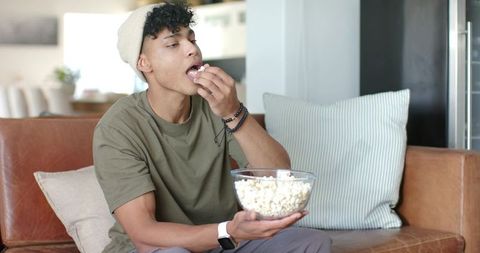 Young man snacking on popcorn on couch in modern open-plan living room, casual leisure