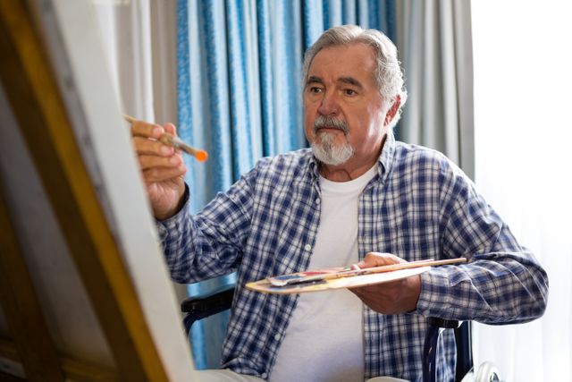 Senior man sitting on a wheelchair, painting on a canvas with a brush and palette in hand. Ideal for use in articles or advertisements related to elderly care, creative hobbies for seniors, nursing home activities, and promoting independence and mental well-being in retirement.