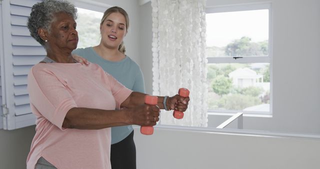 Senior woman practicing strength training with small dumbbells at home, guided by a supportive caregiver. Ideal for showcasing senior fitness, physical therapy, home workouts, and caregiver support. Could be used on websites promoting health, fitness, elderly care, and physical therapy services.