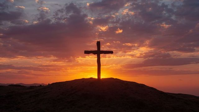 Wooden cross silhouette at sunrise on hilltop with dramatic cloud rays and glowing horizon