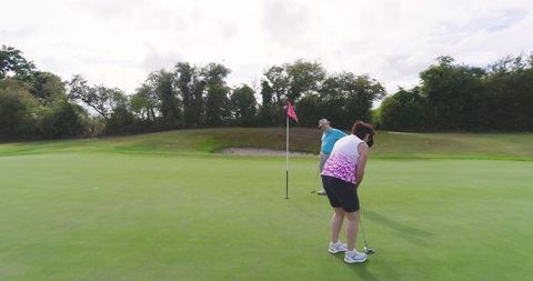 Mature couple playing golf on green, woman putting while senior man watching by flagstick
