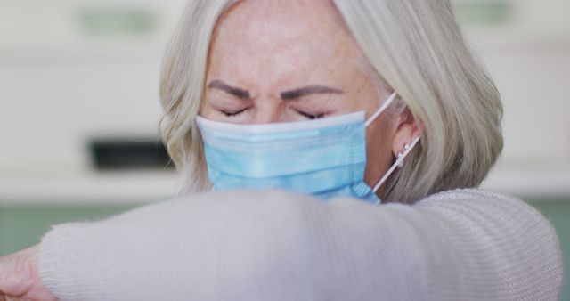 Elderly woman wearing a protective face mask sneezing into her elbow, demonstrating proper hygienic practices during a pandemic. This can be used for health and safety campaigns, illustrating Covid-19 precautions, public health awareness materials, and articles on virus prevention measures.