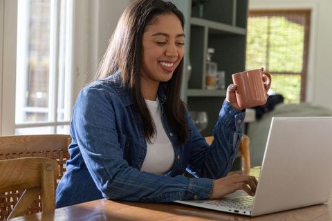 Asian woman working from home on laptop smiling while holding terracotta mug