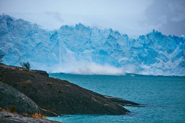 Glacier calving into an icy turquoise ocean bay, creating a splash as ice chunks fall. Rocky shore in the foreground adds to the natural beauty and rawness of the scene. Perfect for topics related to climate change, global warming, and natural splendour.