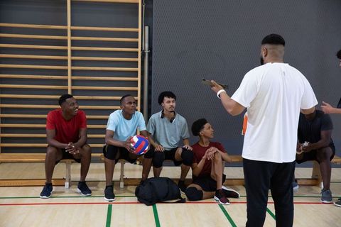 Diverse male volleyball team listening to coach holding tablet on gym bench