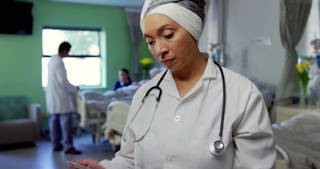 Female nurse wearing head scarf standing in hospital room with stethoscope, looking at patient records. Doctor in background attending to patient. Ideal for illustrating healthcare services, diversity in medical professions, patient care scenes, and modern medical facilities.