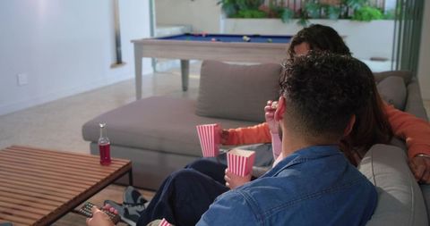 Diverse friends relaxing on gray sofa holding popcorn tubs and remote, casual movie night at home