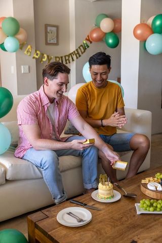 Two men celebrating birthday at home with cake, gifts and balloons on cozy sofa