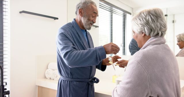Elderly man and woman in bathroom wearing robes, sharing an intimate moment putting toothpaste on a toothbrush. Great for use in healthcare, dental hygiene, senior lifestyle magazines, retirement community advertisements, and articles on healthy aging and daily routines.