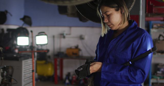Female auto mechanic in a blue uniform, adjusting gloves and preparing for work. Background includes garage lights, tools, and equipment providing an authentic automotive repair scope. Ideal for use in articles or advertisements surrounding gender inclusion in technical fields, automotive service promotions, expert industrial occupations, and skill training programs.