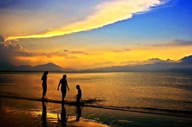 Family walking and playing at beach during sunset with mountains in background. This can be used for advertisements promoting family vacations, travel blogs about beach destinations, or inspirational websites focusing on nature and bonding moments.