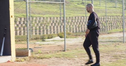 Baseball coach walking field carrying chest protector near dugout with fence and copy space