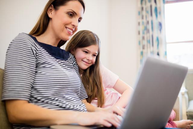 Mother and daughter sitting together on sofa, enjoying time using laptop. Ideal for depicting family bonding, home life, and technology use. Perfect for advertisements or articles focused on family togetherness, modern parenting, or educational technology at home.