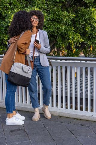 Friends hugging on urban walkway by white railing with phones and crossbody bags