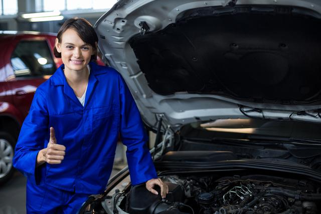 Female mechanic in blue coveralls showing thumbs up while standing beside open car hood in auto repair garage. Ideal for use in automotive industry promotions, workshops, and articles focusing on women in trades or gender diversity in the workplace.