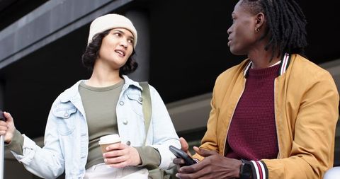 Diverse friends chatting and sipping coffee at modern building entrance, urban casual duo