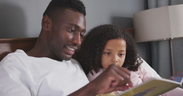 Happy african american father and daughter sitting up in bed reading story book. Fatherhood, childhood, care, togetherness and domestic life.