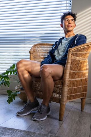 Sunlit asian man sitting in rattan armchair wearing navy hoodie and sneakers, blinds casting stripes