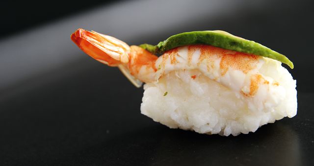 A single piece of sushi featuring a succulent shrimp and a vibrant green avocado slice rests elegantly on a sleek black surface. Its presentation highlights the simplicity and artistry of Japanese cuisine, inviting a taste of the delicate flavors.
