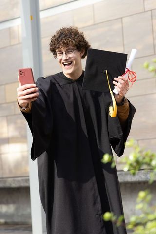 Male graduate celebrating with diploma and smartphone taking selfie in graduation gown