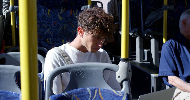 Young man with curly hair concentrating while using a laptop on a bus during daytime. Ideal for illustrating commuting lifestyles, remote work, or studying while traveling. Useful for articles or advertisements regarding public transportation, digital nomadism, or student life.