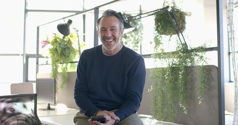 Smiling middle-aged man sitting on desk edge holding smartphone in bright modern office with plants