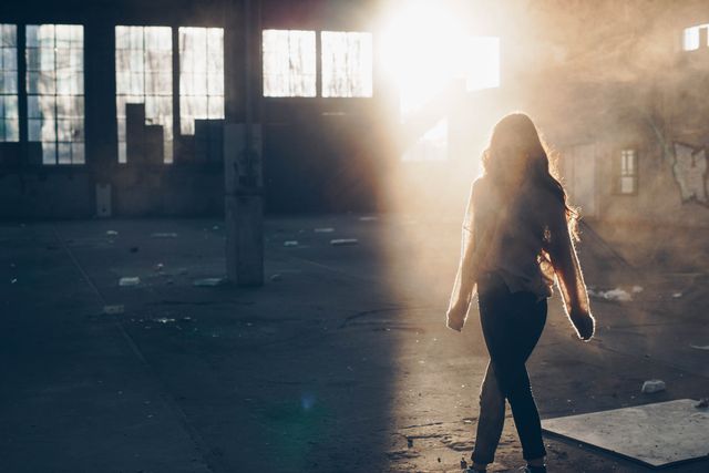 This stock photo depicts a person silhouetted by the setting sun as they walk through an abandoned warehouse. The dust and debris on the floor, combined with the dramatic lighting from the sun's rays, create a mysterious and moody atmosphere. Ideal for use in themes related to urban exploration, solitude, risk-taking adventures, or artistic projects needing a dramatic and intense backdrop.