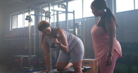 Two mid-adult women performing one-arm dumbbell row with trainer in industrial gym